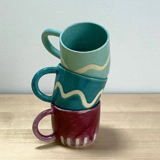 Mug de cerámica Hecho por Su, pintada a mano, personalizada, cerámica artesanal, tres tazas apiladas de colores verde menta, azul y burdeos con diseños ondulados y líneas, sobre superficie de madera.
