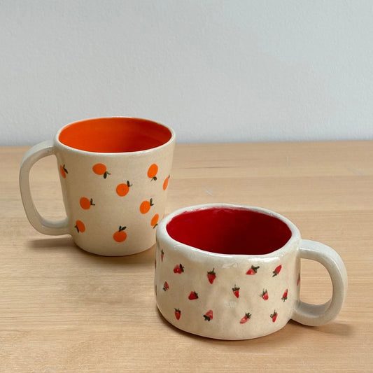 Taza de café Hecho por Su de cerámica artesanal pintada a mano con motivos de naranjas y mug de cerámica personalizada con fresas, interiores naranja y rojo, en mesa de madera.